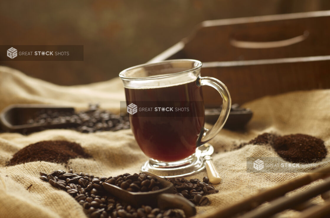Black Coffee in a Glass Mug on a Burlap Material Surface with Coffee Beans and Ground Coffee in an Indoor Setting
