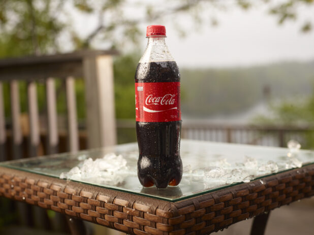 Plastic Bottle of Coca-Cola Brand Cola Soft Drink on a Glass Patio Table with Ice Cubes in an Outdoor Setting