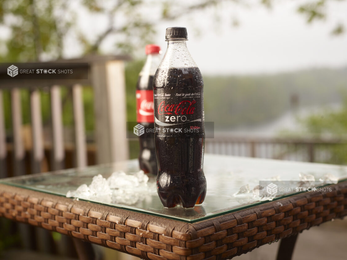 Plastic Bottle of Coca-Cola Brand Coke Zero and Coca-Cola Soft Drinks on a Glass Patio Table with Ice Cubes in an Outdoor Setting