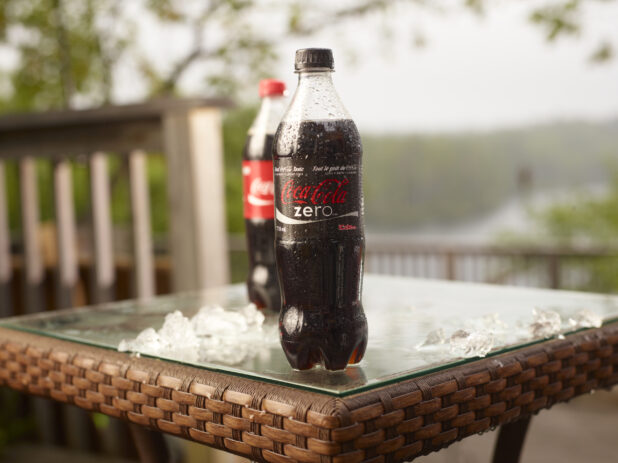 Plastic Bottle of Coca-Cola Brand Coke Zero and Coca-Cola Soft Drinks on a Glass Patio Table with Ice Cubes in an Outdoor Setting