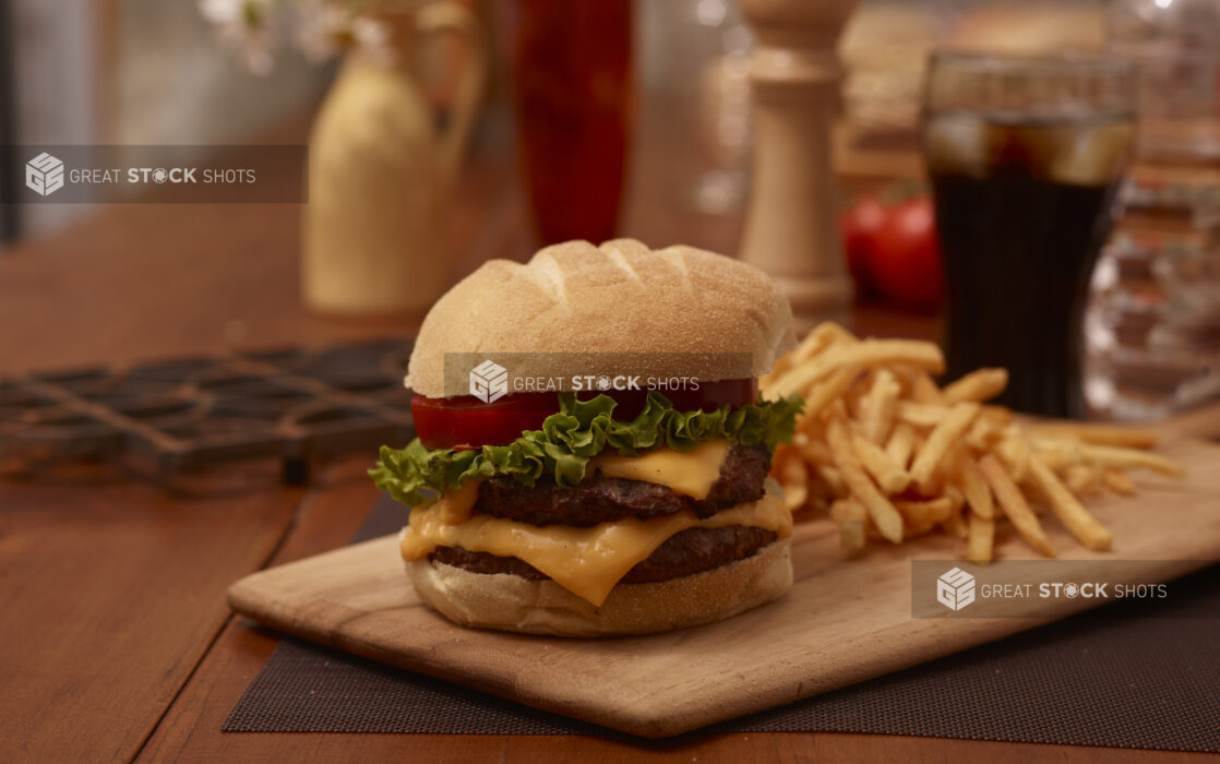 Double Cheeseburger with Lettuce and Tomatoes and a Side of French Fries on a Wooden Platter with a Glass of Soda on a Wooden Table in an Indoor Setting