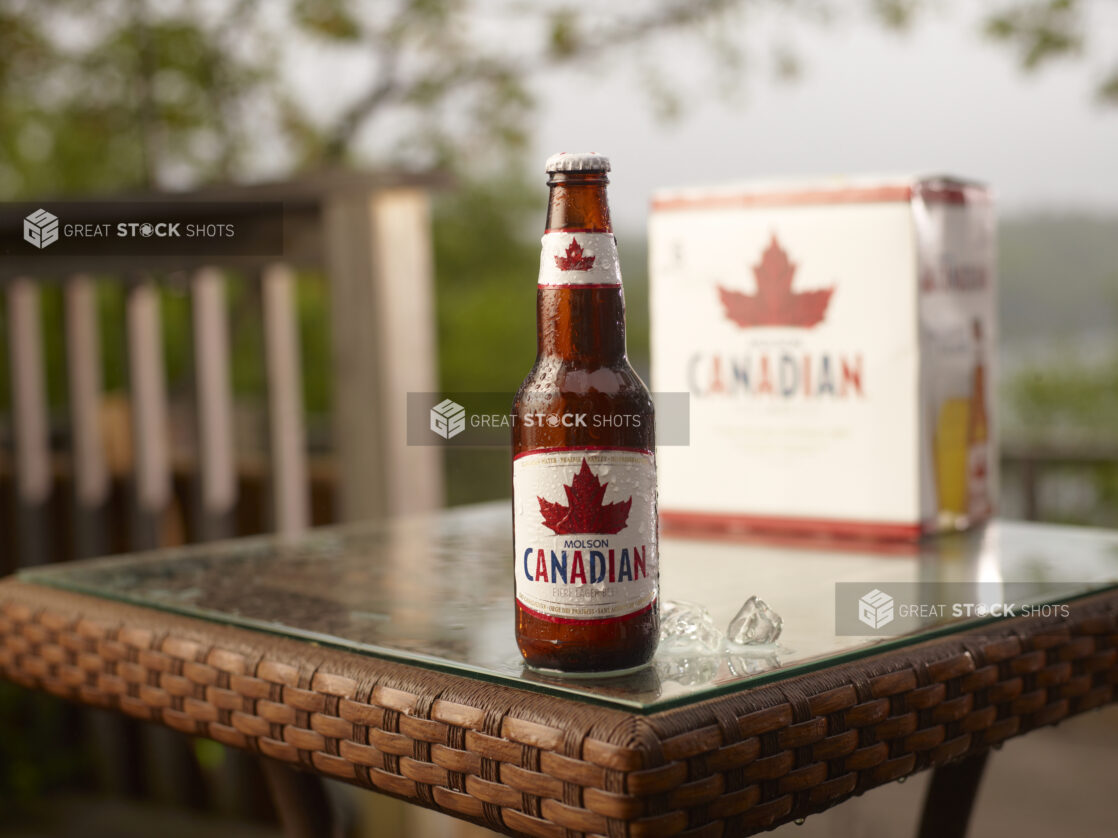 Brown Glass Bottle of Molson Canadian Beer on a Patio Table with a 6-Pack of Bottled Molson Canadian Beer in an Outdoor Setting