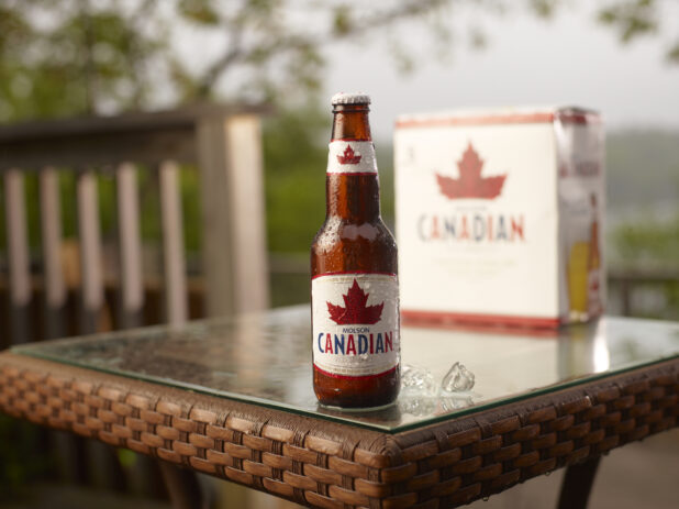 Brown Glass Bottle of Molson Canadian Beer on a Patio Table with a 6-Pack of Bottled Molson Canadian Beer in an Outdoor Setting