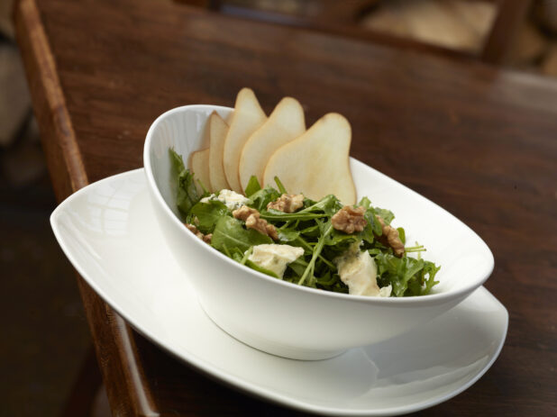 A Bosc Pear, Walnuts, Goat Cheese and Arugula Salad in a Large White Ceramic Bowl on a Dark Wood Surface in a Restaurant Setting