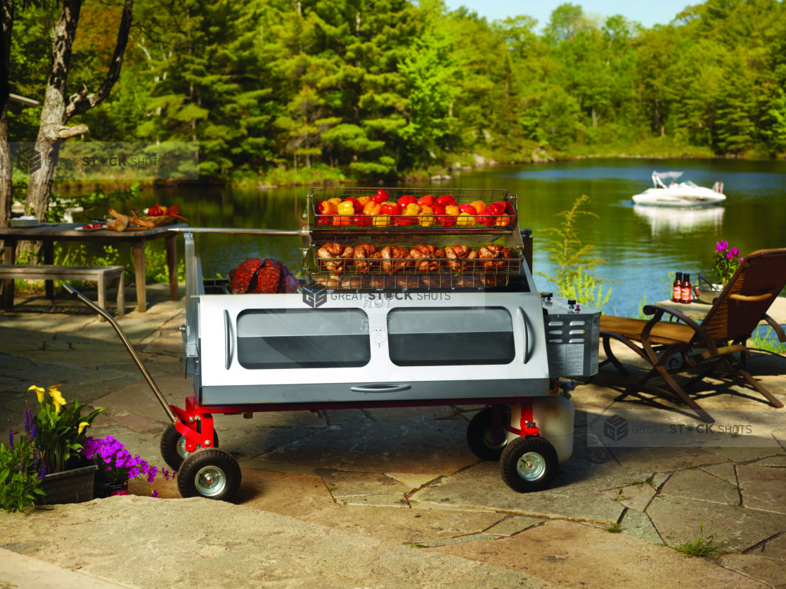 Whole bell peppers and chickens roasting over a propane roaster on a stone terrace with pond and woods in background