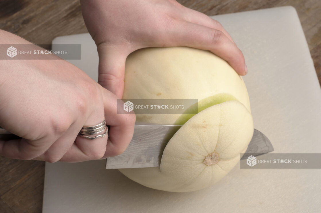 Hands cutting the top from a honeydew melon on a white cutting board on a rustic wooden table