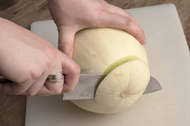 Hands cutting the top from a honeydew melon on a white cutting board on a rustic wooden table
