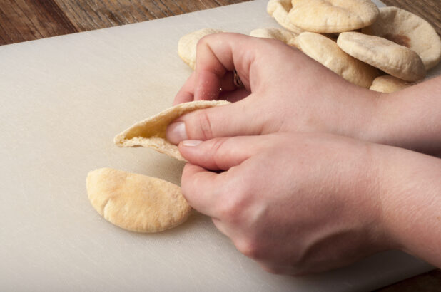 Hands opening a mini pita on a white cutting board on a rustic wooden table