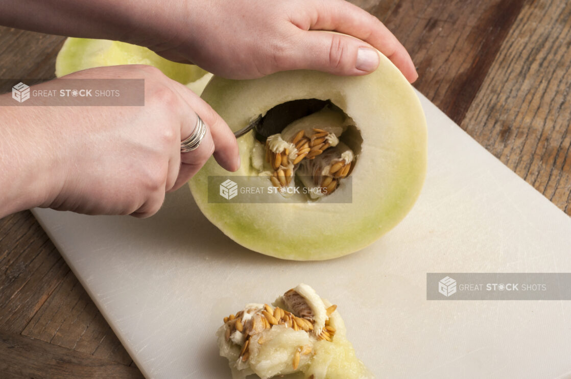 Hands prepping a honeydew melon on a white cutting board on a rustic wooden table