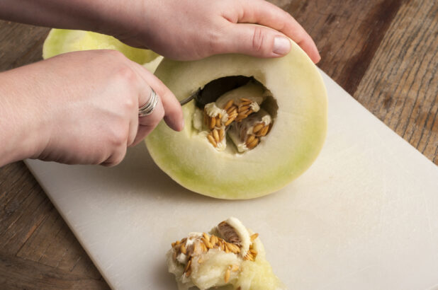 Hands prepping a honeydew melon on a white cutting board on a rustic wooden table