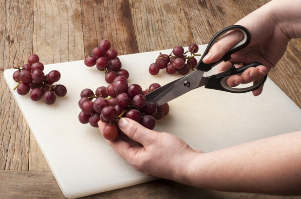 Hands cutting grapes over a white cutting board on a rustic wooden table