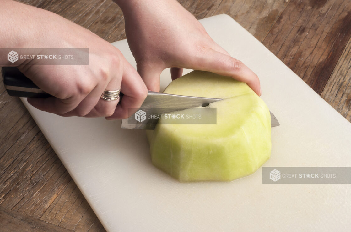 Hands cutting honey dew melon on a white cutting board on a rustic wooden table