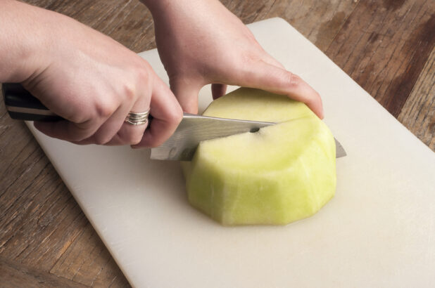 Hands cutting honey dew melon on a white cutting board on a rustic wooden table