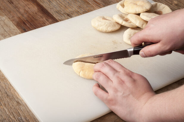 Hands cutting a mini pita with a serrated knife on a white cutting board on a rustic wooden table