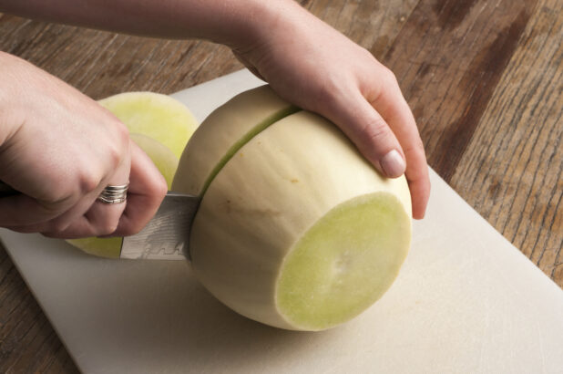 Hands cutting honey dew melon on a white cutting board on a rustic wooden table