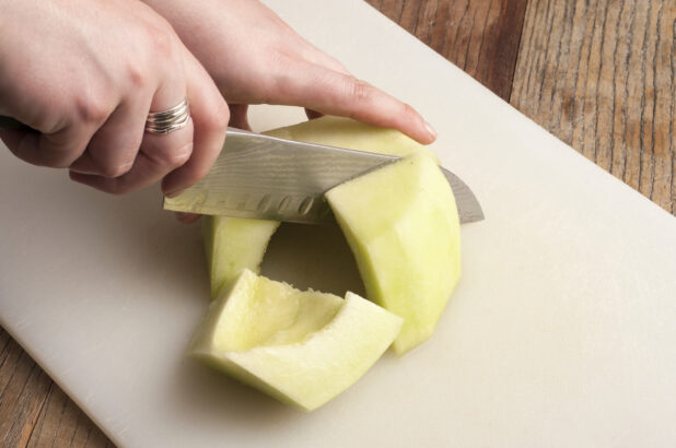 Hands cutting melon on a white cutting board on a rustic wooden table, close-up