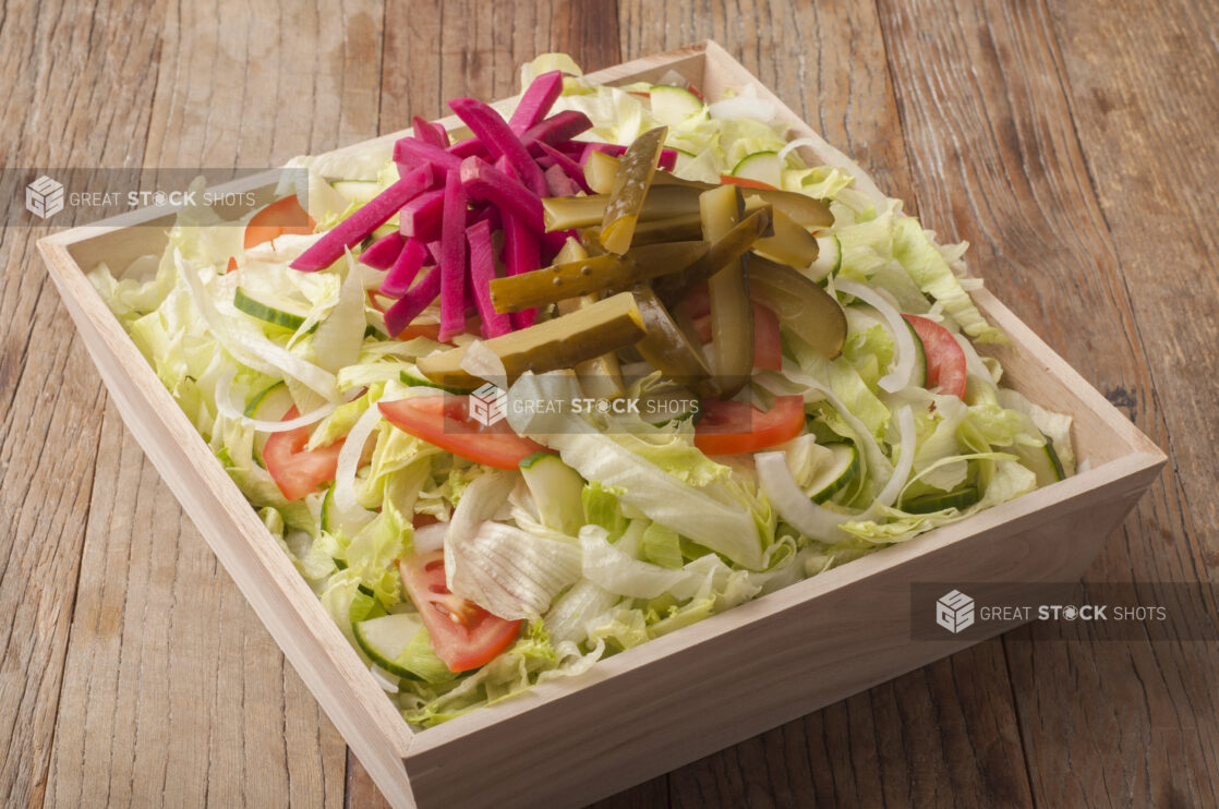 Green salad topped with pickled turnip and cucumbers in a wooden catering box, wood background, close-up