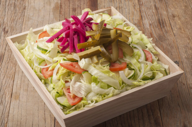 Green salad topped with pickled turnip and cucumbers in a wooden catering box, wood background, close-up