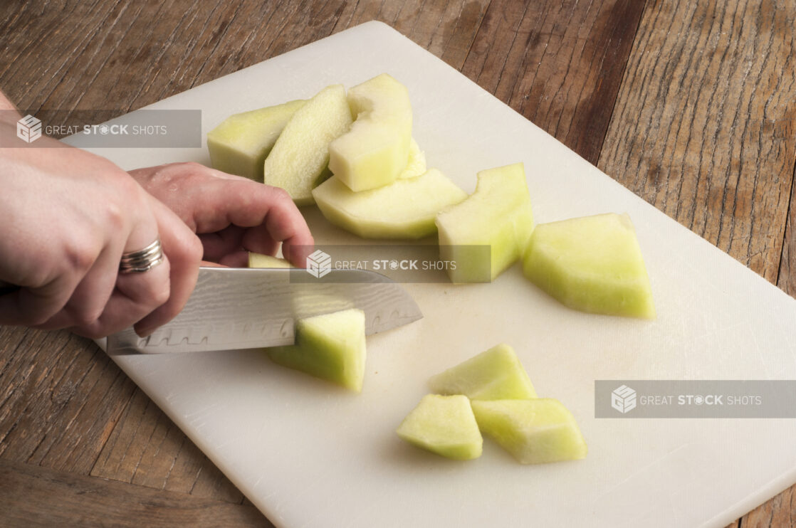 Two hands using a kitchen knife to chop a melon on a white cutting board on a rustic wooden table