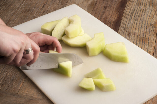 Two hands using a kitchen knife to chop a melon on a white cutting board on a rustic wooden table