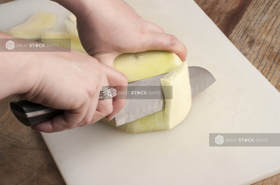 Two hands holding and cutting a melon on a white cutting board on a wooden table with a santoku knife