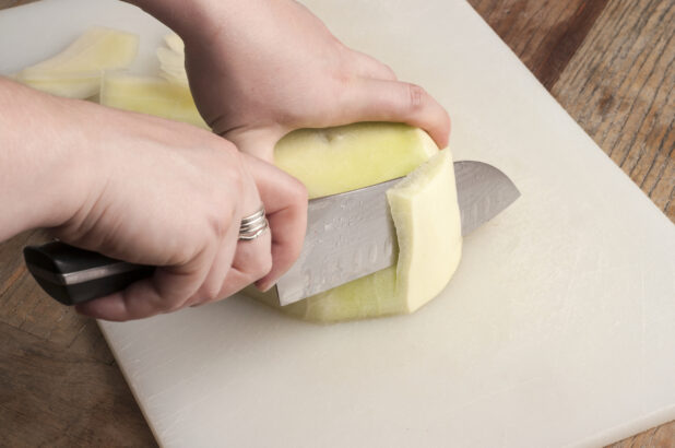 Two hands holding and cutting a melon on a white cutting board on a wooden table with a santoku knife
