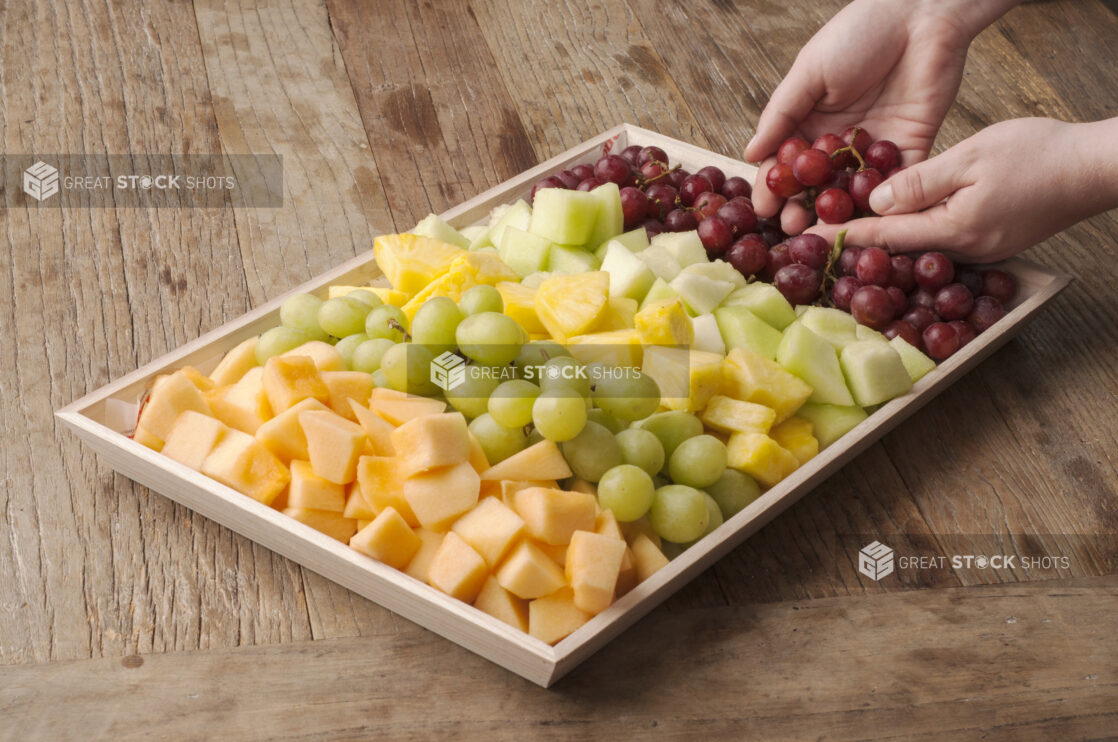 Fruit platter on a wooden tray on a rustic wooden table, hands adding grapes