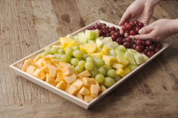 Fruit platter on a wooden tray on a rustic wooden table, hands adding grapes