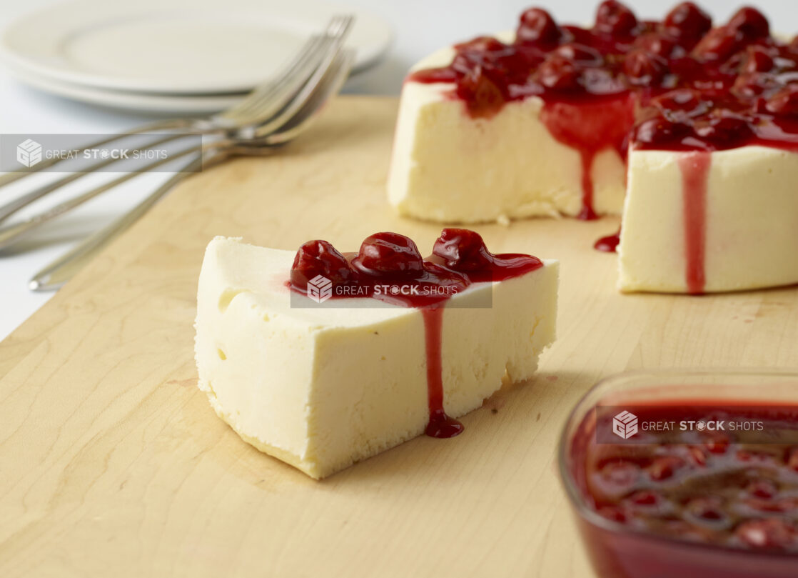 Slice of crustless cheesecake with cherry compote with full cake in the background on a wooden board