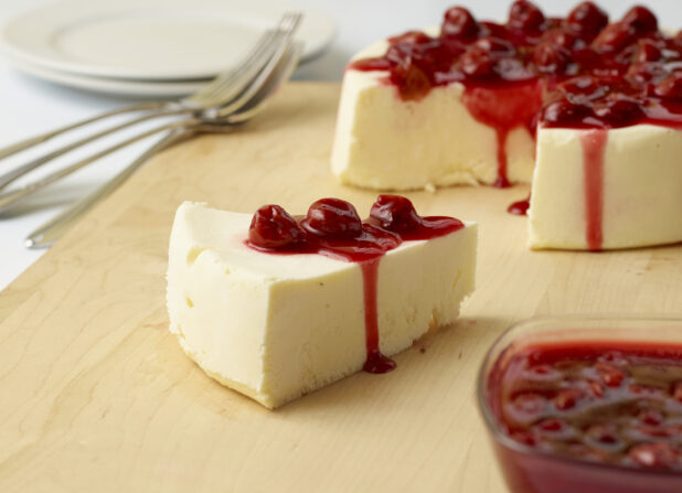 Slice of crustless cheesecake with cherry compote with full cake in the background on a wooden board