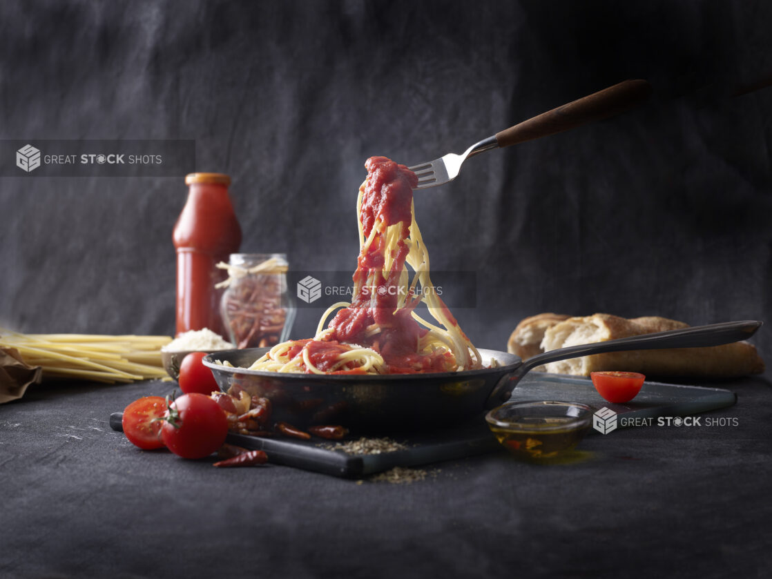 Stainless Steel Saucepan Full of Spaghetti and Marinara Sauce Held Up by a Fork with Campari Tomatoes, Red Chilli and a Baguette Against a Black Backdrop and Surface