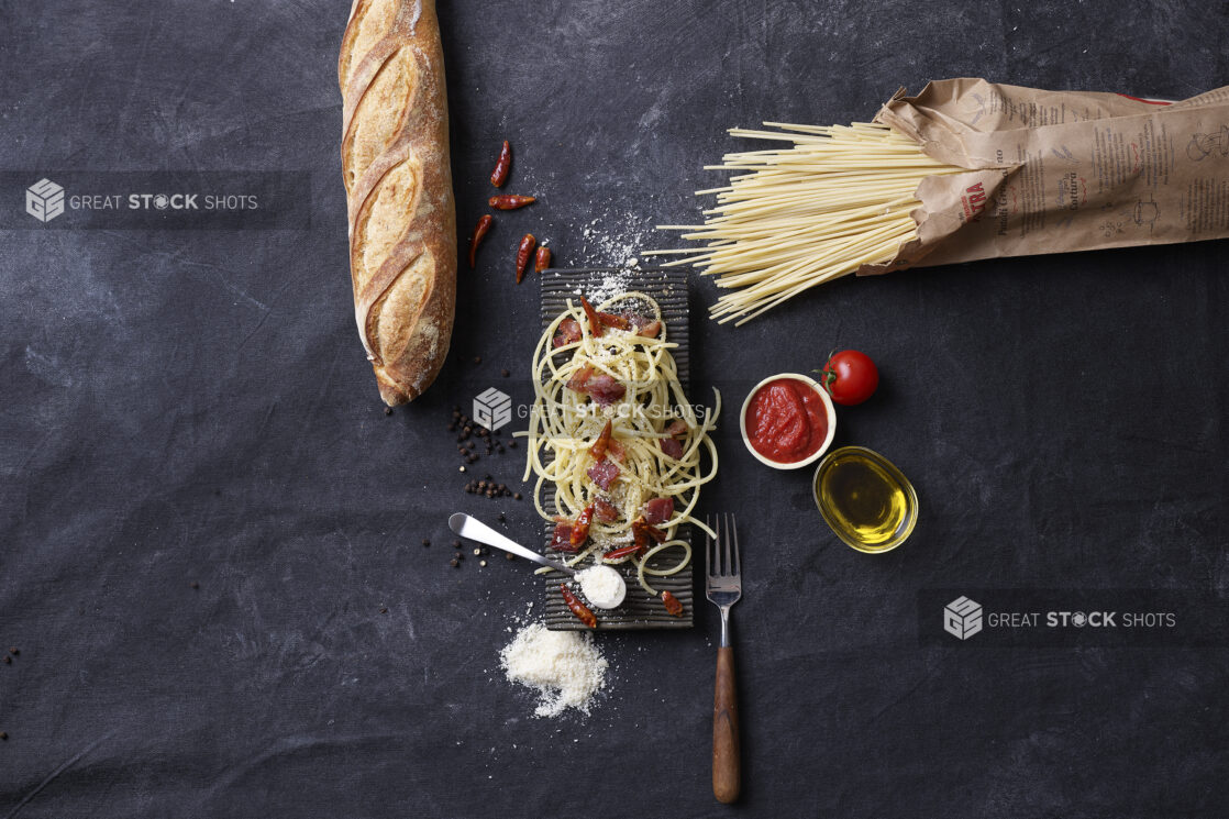 Overhead View of Dried and Cooked Pasta Sun-dried Chillies, Olive Oil and Grated Parmesan Cheese with a Side of Marinara Sauce and a Baguette on a Black Cloth Surface