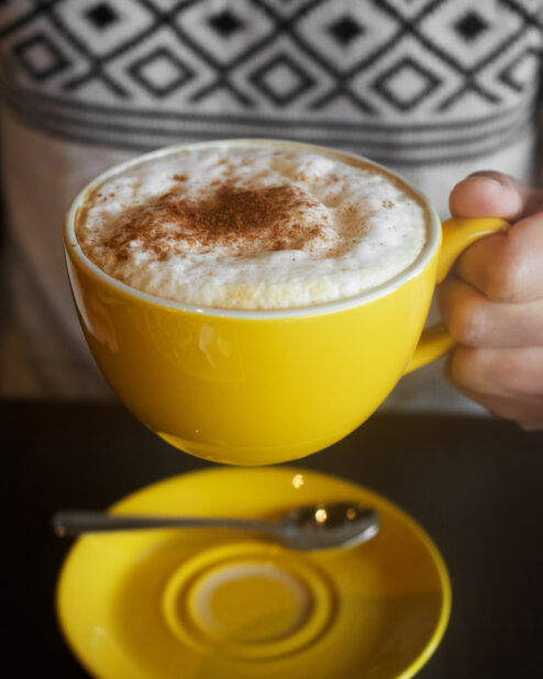 Close Up of a Person Holding a Cappuccino in a Yellow Ceramic Cup with Milk Foam and a Dusting of Cinnamon Powder