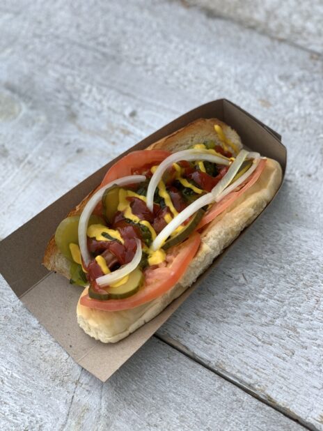 Top down of a hot dog in a cardboard sleeve loaded with tomato, onion, pickles, ketchup, mustard and relish on a wood table