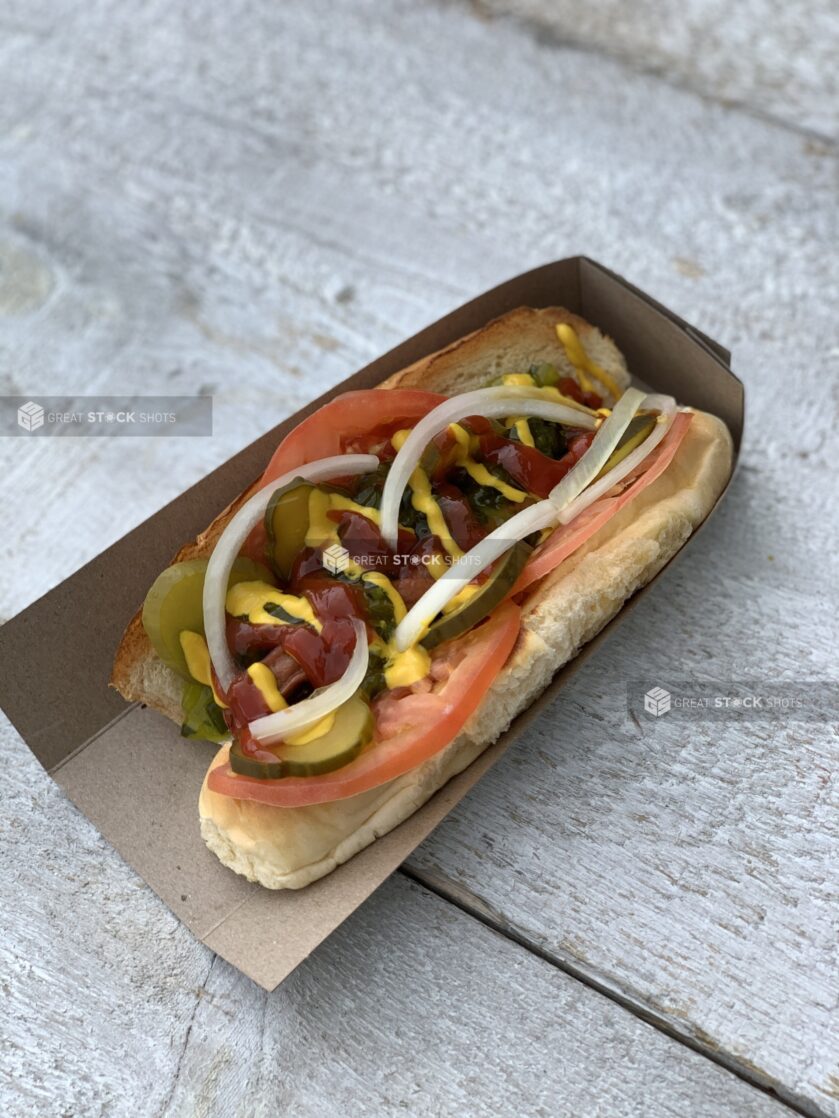 Top down of a hot dog in a cardboard sleeve loaded with tomato, onion, pickles, ketchup, mustard and relish on a wood table