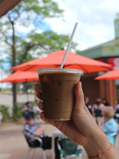 Hand holding up an iced coffee outside on a busy patio with tables and red umbrellas