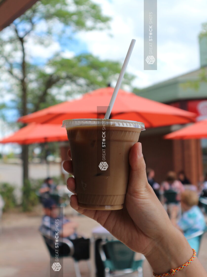 Hand holding up an iced coffee outside on a busy patio with tables and red umbrellas