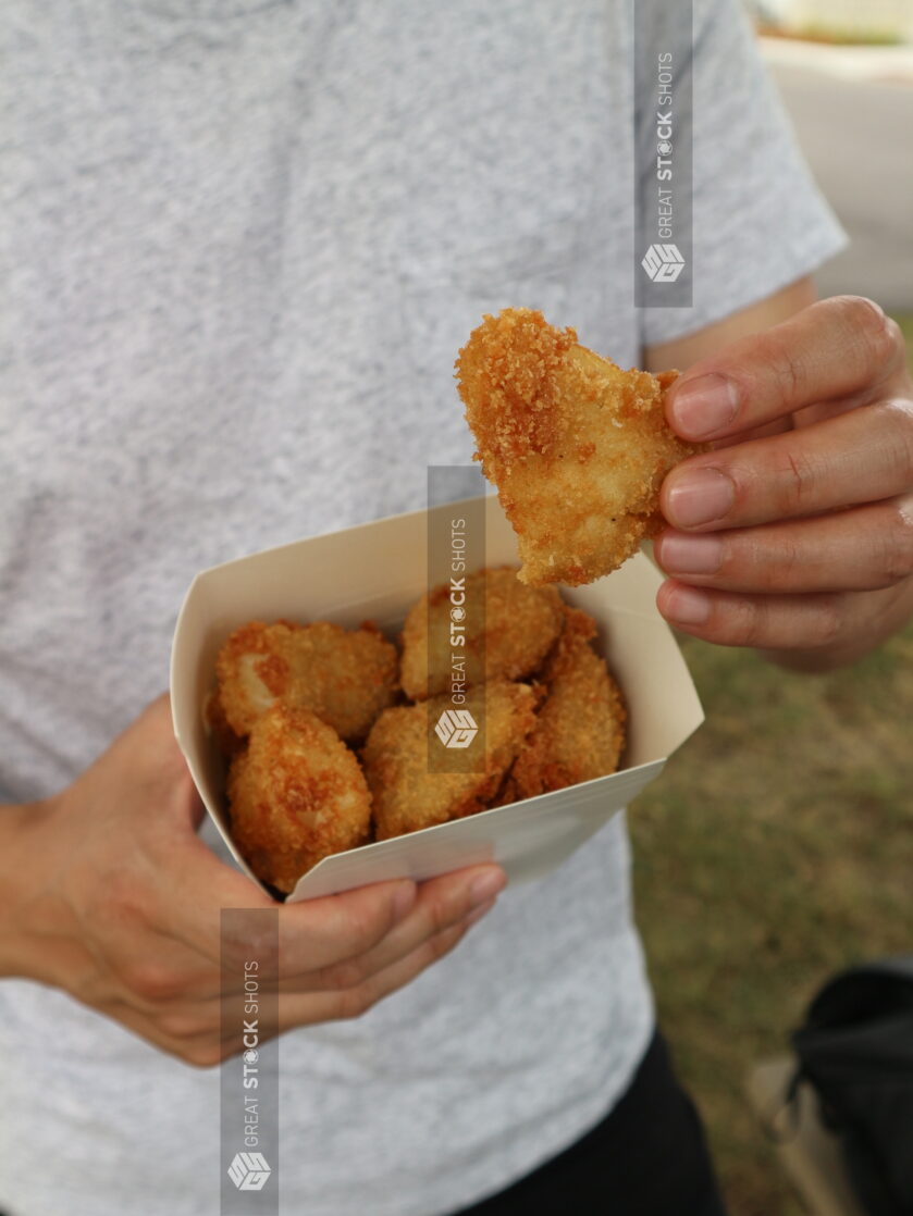 Overhead of person holding a whole chicken nugget over a take out container full of chicken nuggets outside