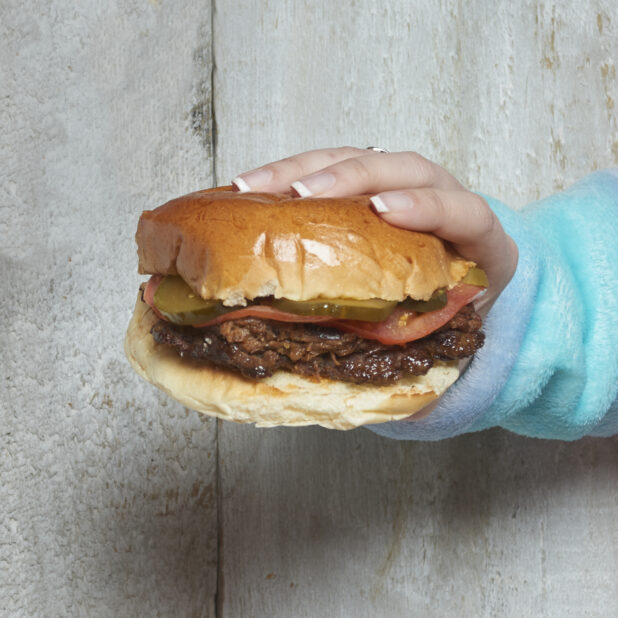 Hand holding a pulled pork sandwich with pickles and tomatoes with wooden background