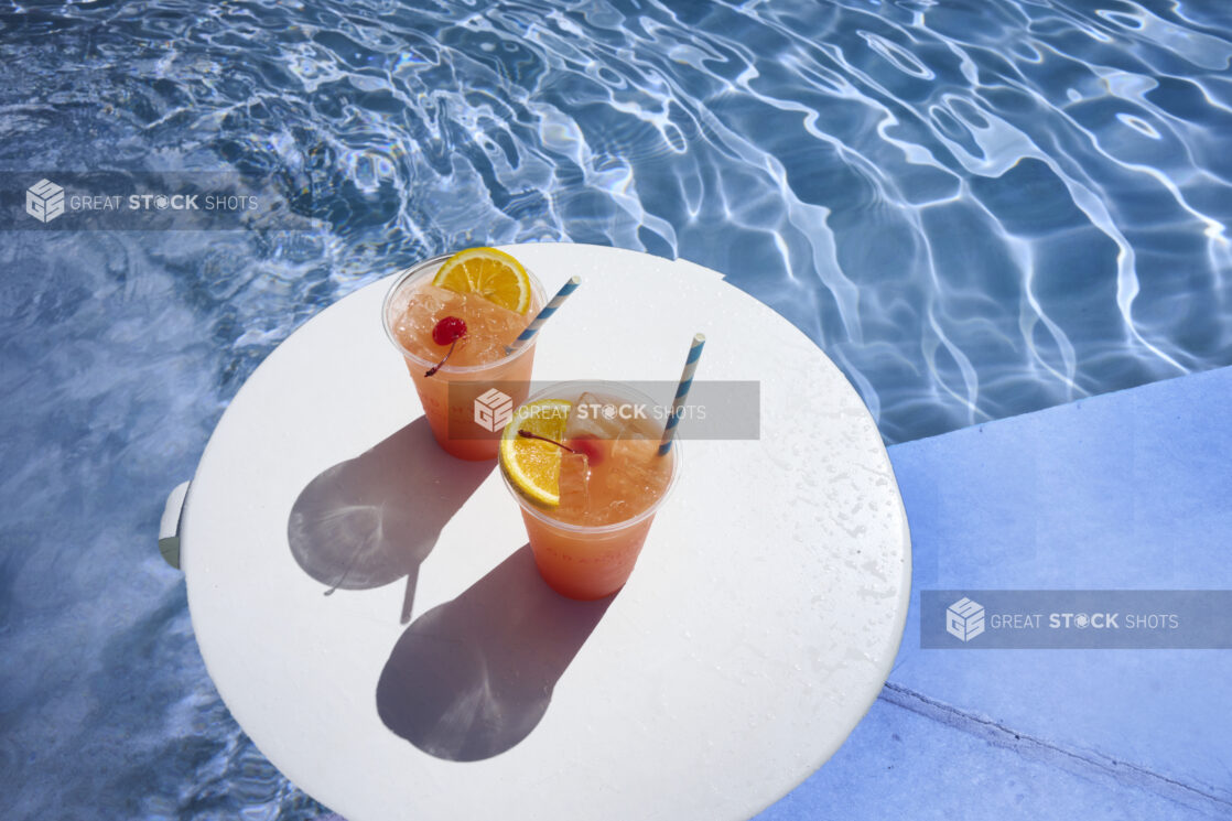 A Pair of Tropical Sunset Cocktails with Cherry and Orange Slice Garnish and Crushed Ice on a Round White Board Floating in an Outdoor Pool