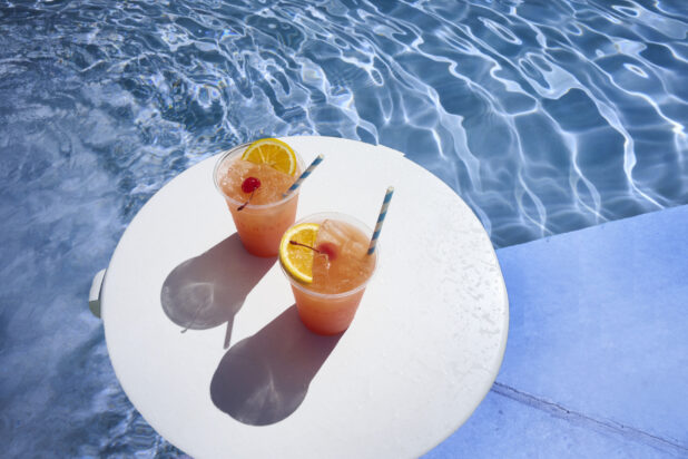 A Pair of Tropical Sunset Cocktails with Cherry and Orange Slice Garnish and Crushed Ice on a Round White Board Floating in an Outdoor Pool