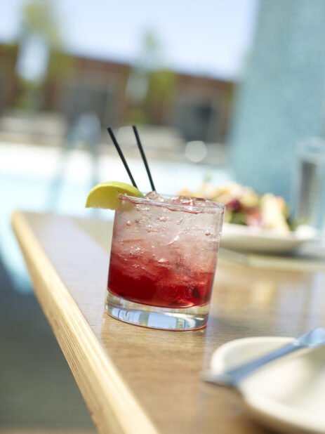 Glass of Cranberry Vodka Cocktail with Crushed Ice and a Lime Wedge on a Wooden Table in an Outdoor Poolside Patio Setting