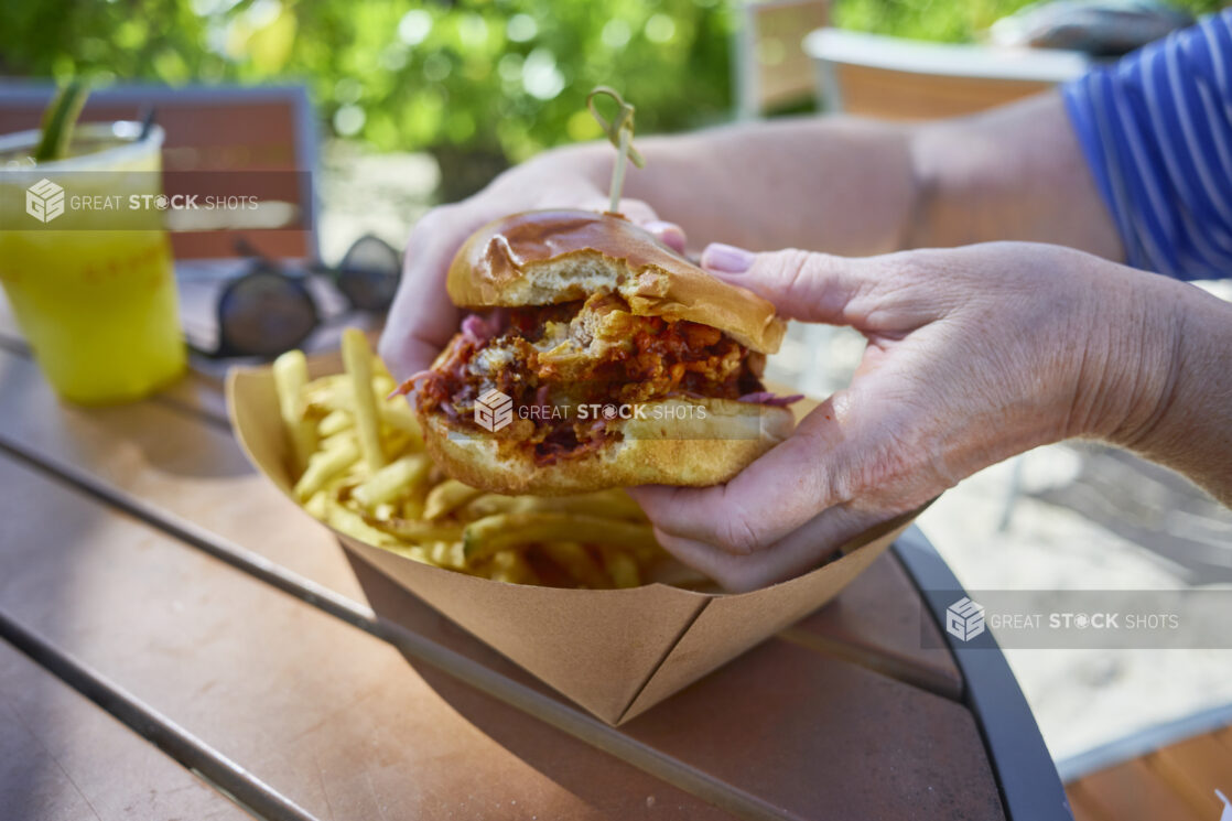 Close-Up of Hands Holding Up a Spicy Chicken Burger With a Bite Taken Out of It With a Take-Out Container of French Fries on a Patio Table Outdoors