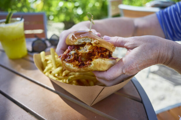 Close-Up of Hands Holding Up a Spicy Chicken Burger With a Bite Taken Out of It With a Take-Out Container of French Fries on a Patio Table Outdoors