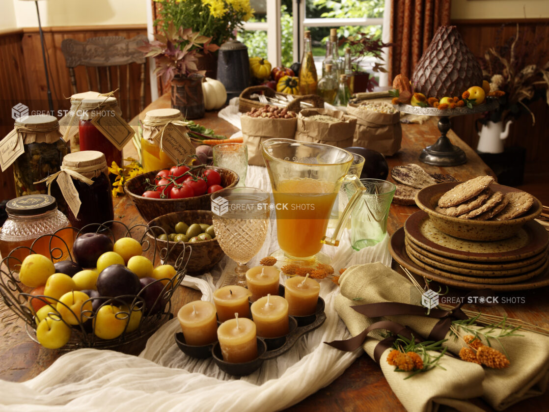 Autumn Table Setting with Homemade Jars of Jam, Pickles, Sauces, Baskets of Fresh Plums and Tomatoes, Fresh Apple Cider, Green Olives and Cookies on a Wooden Table in an Indoor Setting