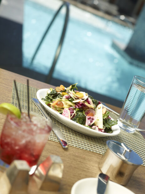 A Large Fresh Salad of Kale, Watermelon Radish, Pears, Grapes and Radicchio in a Round White Ceramic Bowl on a Woven Placemat in a Poolside Patio Setting Outdoors