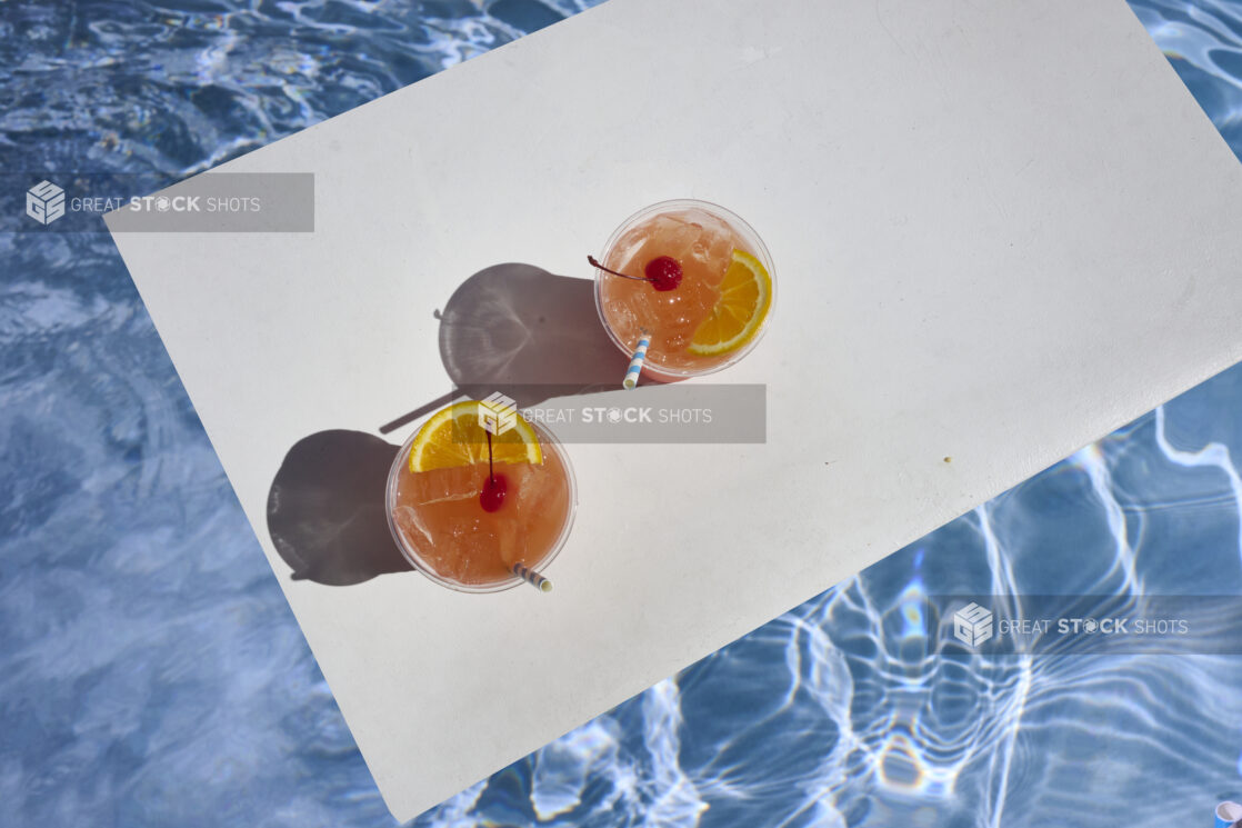 Overhead View of a Pair of Tropical Sunset Cocktails with Cherry and Orange Slice Garnish and Crushed Ice on a White Board Floating in an Outdoor Pool