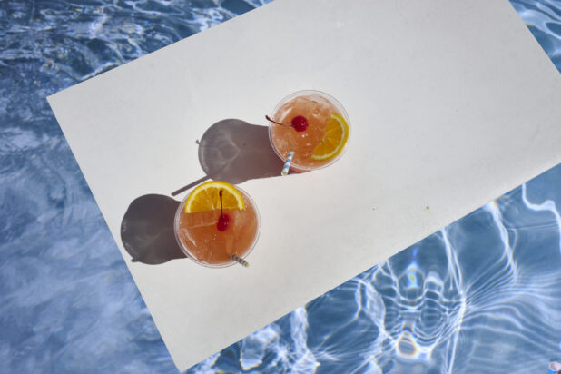 Overhead View of a Pair of Tropical Sunset Cocktails with Cherry and Orange Slice Garnish and Crushed Ice on a White Board Floating in an Outdoor Pool