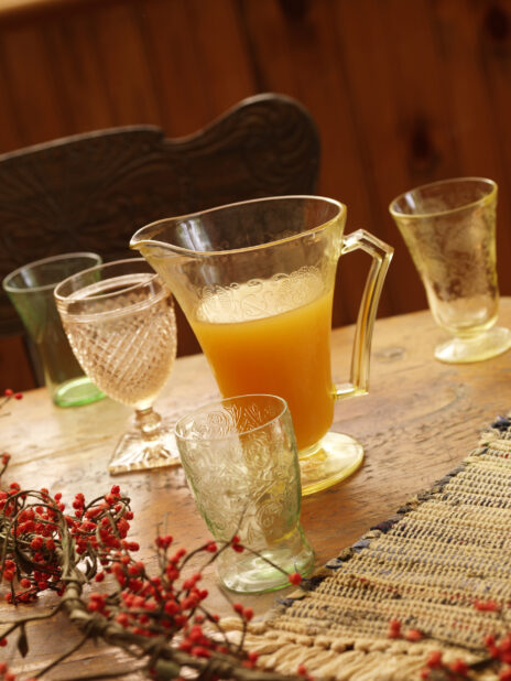 A Glass Jug of Apple Cider on a Wooden Table Surrounded by Assorted Glass Cups with a Woven Placemat and Winter Berries in an Indoor Setting