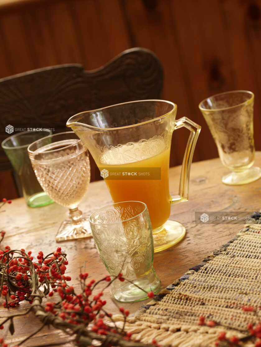 A Glass Jug of Apple Cider on a Wooden Table Surrounded by Assorted Glass Cups with a Woven Placemat and Winter Berries in an Indoor Setting